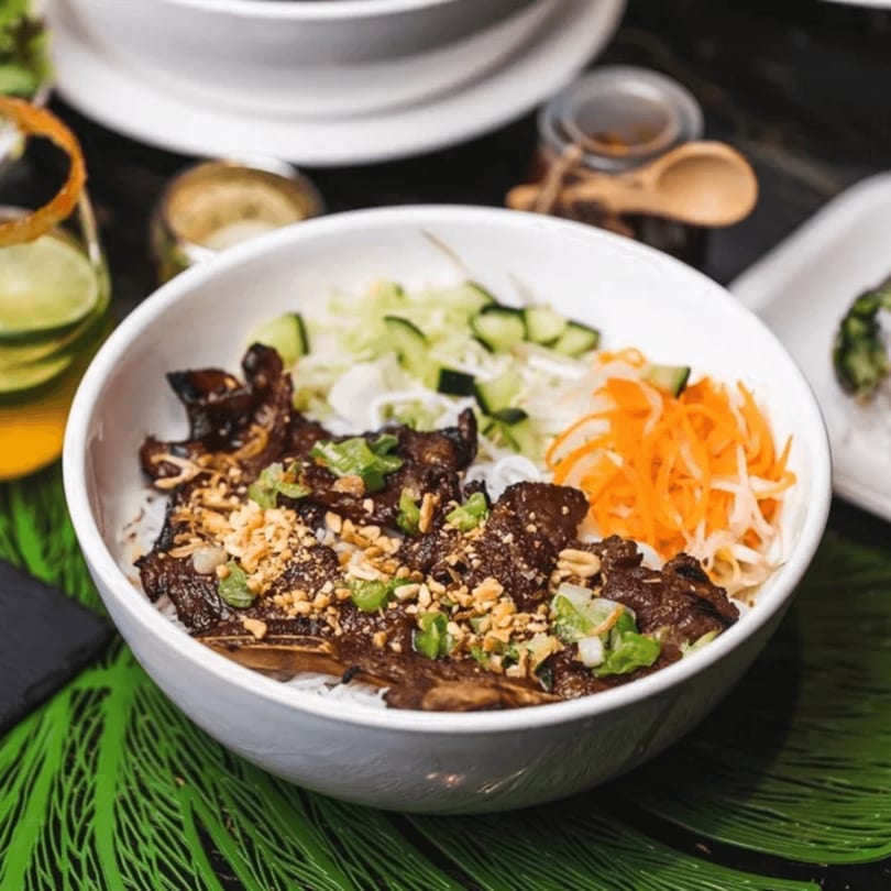 A white bowl of Vietnamese vermicelli noodles with grilled beef, vegetables, and peanuts.