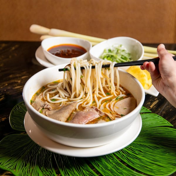 A hand uses chopsticks to lift noodles from a bowl of Vietnamese bun bo hue soup.