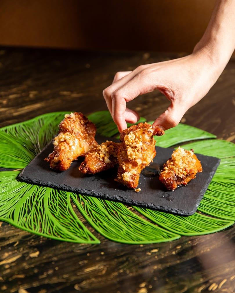 A hand picks up a crispy fried chicken wing topped with peanuts from a slate plate.